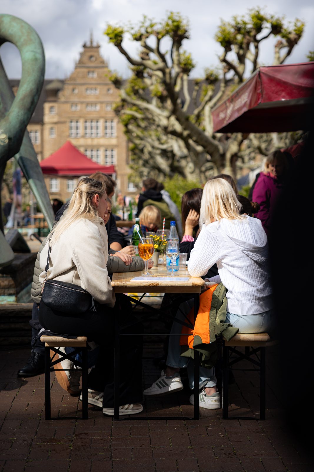Menschen an langen Tischen unter Platanen auf dem Friedensplatz